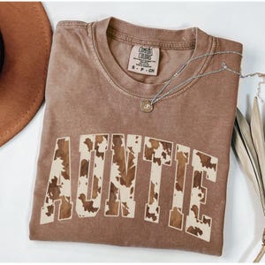 May include: Auntie t-shirt in a brown color with the word "AUNTIE" in a cowhide pattern. The shirt has a crew neck and short sleeves. A silver necklace with a pendant is draped across the shirt. A brown hat is visible in the upper left corner.