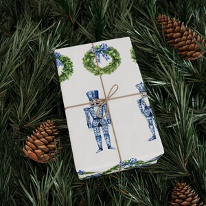 Puede incluir: Una caja de regalo rectangular envuelta en papel blanco con un patr&oacute;n repetitivo de cascanueces azules y blancos y coronas verdes con lazos azules, atada con un cordel marr&oacute;n fino centrado sobre la parte superior del paquete.
