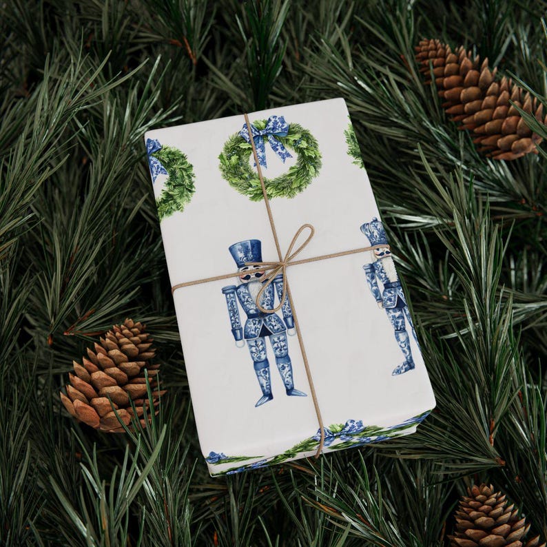 Puede incluir: Una caja de regalo rectangular envuelta en papel blanco con un patr&oacute;n repetitivo de cascanueces azules y blancos y coronas verdes con lazos azules, atada con un cordel marr&oacute;n fino centrado sobre la parte superior del paquete.