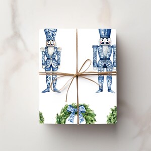 Puede incluir: Una caja de regalo rectangular envuelta en papel blanco con un patr&oacute;n repetitivo de cascanueces azules y blancos y coronas verdes con lazos azules. El paquete est&aacute; atado con un cordel marr&oacute;n fino centrado en un lazo en la parte frontal.