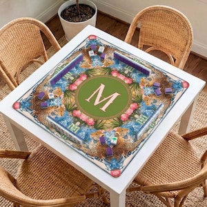 Puede incluir: Una mesa cuadrada blanca con un juego de Mahjong decorativo. El tablero presenta un c&iacute;rculo verde con la letra "M", rodeado de un dise&ntilde;o floral y de leopardo. Sillas de mimbre rodean la mesa.