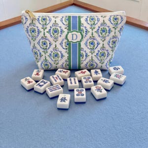 May include: A white cosmetic bag with a blue and green floral pattern and a gold zipper. The bag has a green and blue stripe with a white letter "D" in the center. Scattered in front are white and blue Mahjong tiles.