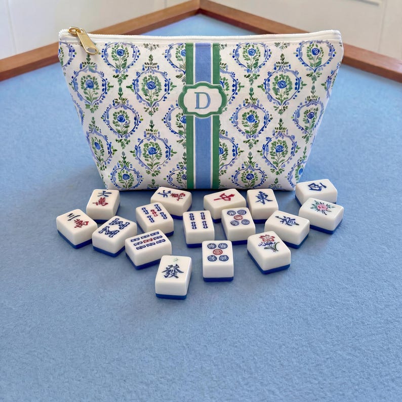 May include: A white cosmetic bag with a blue and green floral pattern and a gold zipper. The bag has a green and blue stripe with a white letter "D" in the center. Scattered in front are white and blue Mahjong tiles.