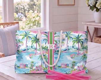 May include: A rectangular tote bag featuring a tropical landscape print with palm trees, pink buildings, and blue water. The bag has a vertical green and pink center stripe with the letter B in a white square and thick white rope handles.