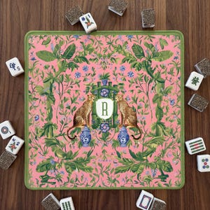 Puede incluir: Un tapete de Mahjong rosa con borde verde, con un diseño floral y de guepardo. El tapete tiene un escudo central con la letra "R" y dos guepardos. Las fichas de Mahjong están esparcidas.