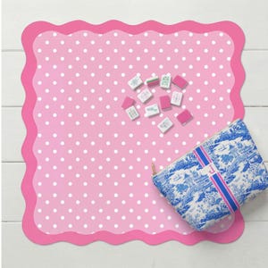 May include: A pink polka-dot placemat with a scalloped edge, featuring a blue and white toiletry bag with a pink stripe and the letter "J". Scattered on the placemat are small, rectangular game tiles.