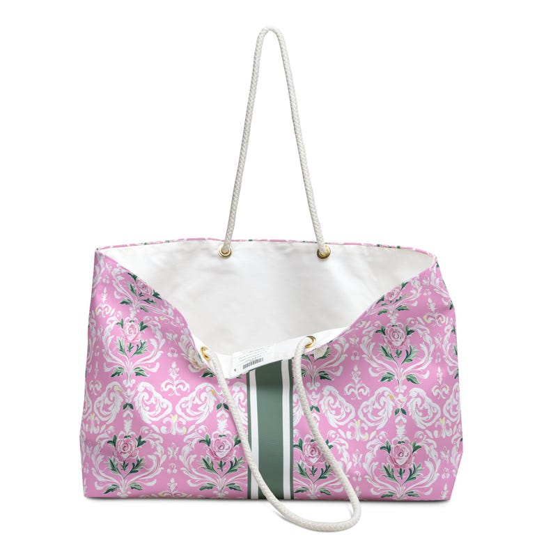 May include: A large tote bag featuring a pink damask pattern with white floral motifs and green accents. The bag has a solid dark green vertical stripe down the center, a white interior lining, and two white braided rope handles with gold grommets.