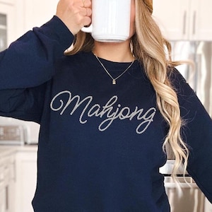 May include: A navy blue long-sleeve sweatshirt featuring the word Mahjong written in a cursive script across the chest. The lettering is composed of small, white, circular beads or pearls that create a textured, raised appearance on the fabric.