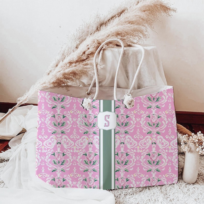 May include: A tote bag featuring a pink damask pattern with green floral accents and a central vertical green stripe. The bag includes a white rectangular emblem with the letter S in pink, and two thick white rope handles attached at the top.