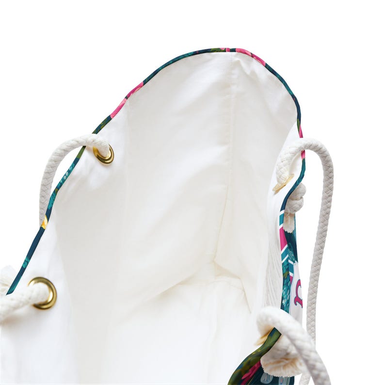 May include: A tote bag with a white interior lining and thick white rope handles attached through gold-toned metal grommets. The exterior features a dark teal background with a vibrant floral pattern in shades of pink, green, and white.