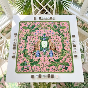 May include: A white square Mahjong table with a pink floral patterned playing surface. The surface features two cheetahs, blue vases, and the letter "R". Green trim surrounds the playing area. Mahjong tiles are arranged around the table.