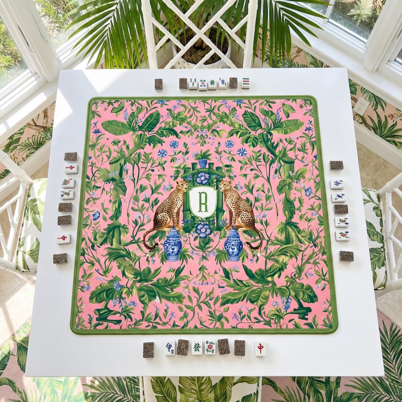 May include: A white square Mahjong table with a pink floral patterned playing surface. The surface features two cheetahs, blue vases, and the letter "R". Green trim surrounds the playing area. Mahjong tiles are arranged around the table.