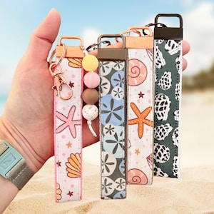 May include: A hand holds four fabric key fobs with various beach-themed patterns. The patterns include seashells, starfish, and floral designs in pink, blue, and green. Each fob has a metal clasp and a wrist strap. Some have decorative beads.