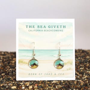 May include: A pair of silver-toned drop earrings featuring circular pendants with a teal and brown mountain landscape design. The earrings are displayed on a white card printed with the text THE SEA GIVETH CALIFORNIA BEACHCOMBING BORN OF SAND & SEA.