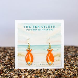 May include: A pair of orange fish-shaped earrings with gold-colored hooks. The earrings are displayed on a white card with the text "THE SEA GIVETH CALIFORNIA BEACHCOMBING" and a beach scene illustration. The earrings are set against a background of small pebbles.