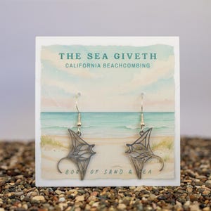 以下が含まれることがあります： 金色のフックに吊るされた、マンタの形をした銀色のイヤリングのペア。 イヤリングは、「THE SEA GIVETH CALIFORNIA BEACHCOMBING」と「BORN OF SAND & SEA」のテキストが水彩画のビーチシーンの上に表示されたカードに表示されています。