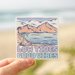 May include: A sticker with a beach scene featuring mountains, palm trees, and a surfboard. The text "LOW TIDES GOOD VIBES" is displayed in a gradient font. The sticker is held by a hand.