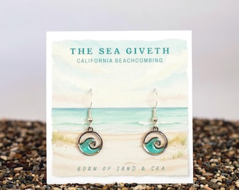 May include: Silver dangle earrings with a circular design featuring a turquoise wave. The earrings are displayed on a card with the text "THE SEA GIVETH CALIFORNIA BEACHCOMBING" and "BORN OF SAND & SEA" against a beach scene backdrop.