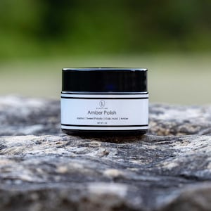 May include: A small, white jar with a black lid and label. The label reads "Amber Polish" and lists the ingredients: Jojoba, Sweet Potato, Kojic Acid, Amber. The jar is sitting on a gray rock.