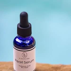 May include: A blue glass bottle with a black dropper top and a white label. The label reads "B. Skincare Pearl Serum Hyaluronic Acid B5 | Pearl Net 1 Fl Oz".