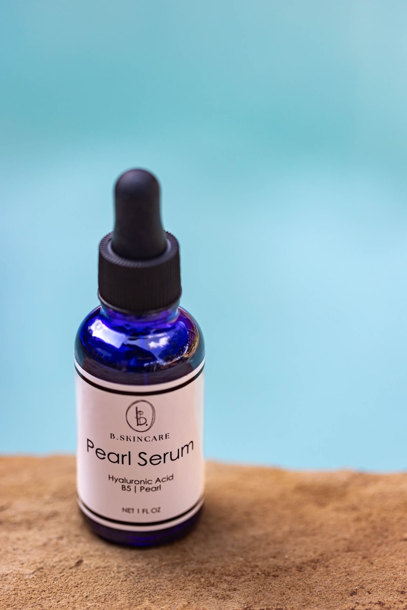 May include: A blue glass bottle with a black dropper top and a white label. The label reads "B. Skincare Pearl Serum Hyaluronic Acid B5 | Pearl Net 1 Fl Oz".