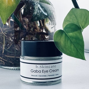 May include: A white jar with a black lid and a label that reads "b.Skincare Gaba Eye Cream Broccoli | Ceramides | Periwinkle | Gaba"
