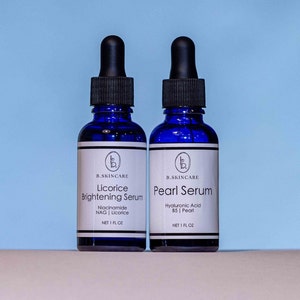 May include: Two blue glass bottles with dropper tops containing skincare serums. The bottle on the left is labeled "Licorice Brightening Serum" with the ingredients "Niacinamide NAG | Licorice" and "NET 1 FL OZ". The bottle on the right is labeled "Pearl Serum" with the ingredients "Hyaluronic Acid B5 | Pearl" and "NET 1 FL OZ".