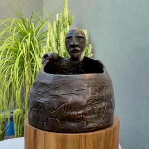 Puede incluir: Una maceta de madera tallada de color marrón oscuro con una figura humana que emerge de la parte superior. La maceta tiene diseños grabados y se asienta sobre una base de madera natural. Plantas verdes y cactus en el fondo.