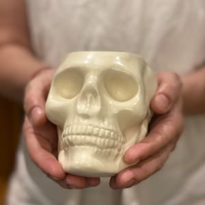 May include: A cream-colored skull-shaped ceramic container. The skull has detailed facial features, including eye sockets, a nose cavity, and teeth. The container is held in front of a soft, blurred background.