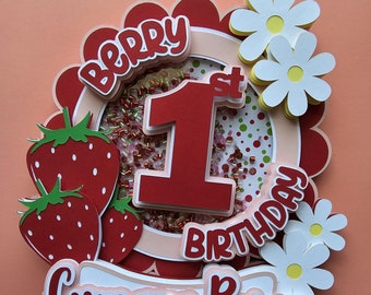 May include: A red and white cake topper with the words "Berry 1st Birthday" and "Cymara Reign". The topper features a large red number 1, strawberries, and white daisies.