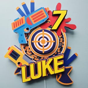 May include: A colorful cake topper featuring a blue and orange toy gun, a target, and the name "LUKE" in yellow letters. A large yellow number 7 is also included, with red accents. The topper is designed for a birthday celebration.