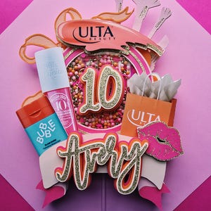 May include: A layered paper cake topper featuring the name Avery in gold glitter script, the number 10, an Ulta Beauty logo, a miniature orange shopping bag, a pink lip icon, and cutouts of beauty products including a Sol de Janeiro bottle.