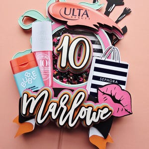 May include: A layered paper cake topper with the name "Marlowe" in white script. The topper includes a "10" with black and white accents, a "Sephora" shopping bag, and a pink lip design. Other elements include "Ulta Beauty" and beauty product replicas.