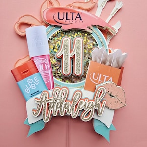 May include: A decorative cake topper with the Ulta Beauty logo, the number 11, and the name "Addleigh" in gold glitter. The topper includes miniature cosmetic products and a shopping bag, all set against a pink background.