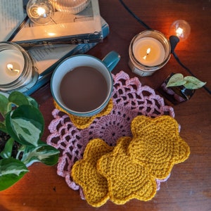 Puede incluir: Primer plano de un encaje de ganchillo con tres posavasos de ganchillo en forma de estrella de color amarillo mostaza. El encaje es rosa y blanco. Una taza de café se encuentra sobre un posavasos y hay dos velas en el fondo.