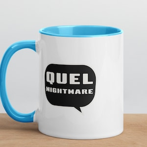 Puede incluir: Taza de cerámica blanca con borde y asa de color azul claro. La taza tiene una burbuja de diálogo negra con el texto "QUEL NIGHTMARE" en blanco.