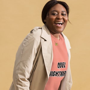 Puede incluir: Una mujer lleva una sudadera de color melocotón con el texto "QUEL NIGHTMAR" en letras negras que gotean. Lleva una gabardina beige y sonríe.