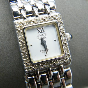 May include: A silver Anne Klein wristwatch with a rectangular face. The watch has a white dial with black hands and Roman numeral hour markers. The bezel and strap are decorated with small, sparkling stones. The watch reads "Diamond Swiss".