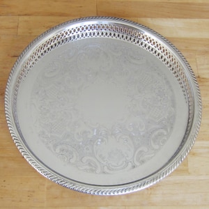May include: A round silver-toned serving tray featuring an intricate engraved floral pattern in the centre and a decorative pierced lattice border. The rim of the tray is finished with a twisted rope-style edge, providing a textured metallic appearance.