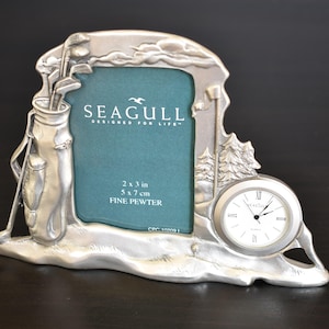 May include: Silver pewter golf-themed photo frame with a clock. The frame holds a 5 x 7 cm photo and features a golf bag, clubs, and a clock with Roman numerals. The text "SEAGULL" is visible.