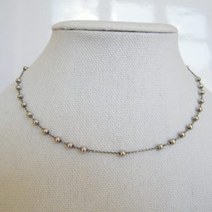 Può includere: Una delicata collana a catena color argento caratterizzata da piccole sfere metalliche distanziate uniformemente lungo tutta la sua lunghezza. La collana è progettata come un semplice filo continuo di perline collegate da sottili maglie metalliche.