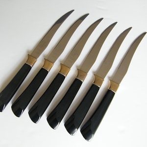 May include: A set of six steak knives. Each knife has a serrated silver blade, a black handle, and a gold-coloured bolster. The knives are arranged on a white surface.