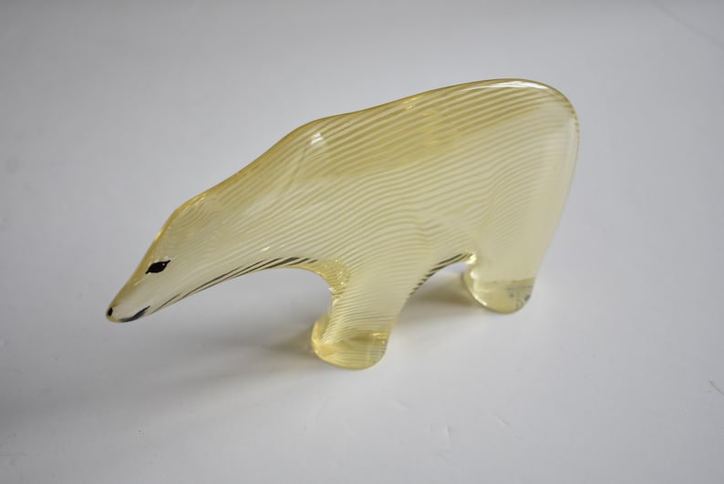 K&ouml;nnte beinhalten: Eine gelbe Glasfigur eines Eisb&auml;ren mit einem gestreiften Muster. Die Figur steht auf einer wei&szlig;en Oberfl&auml;che.