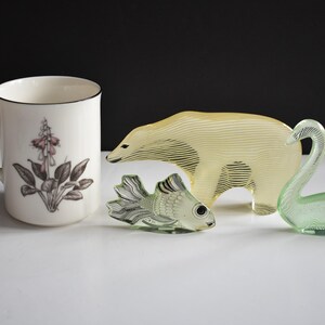 K&ouml;nnte beinhalten: Eine wei&szlig;e Keramiktasse mit Blumenmuster, eine gelbe Glasfigur eines Eisb&auml;ren, eine gr&uuml;ne Glasfigur eines Schwans und eine gr&uuml;ne Glasfigur eines Fisches.