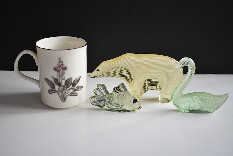 K&ouml;nnte beinhalten: Eine wei&szlig;e Keramiktasse mit Blumenmuster, eine gelbe Glasfigur eines Eisb&auml;ren, eine gr&uuml;ne Glasfigur eines Schwans und eine gr&uuml;ne Glasfigur eines Fisches.