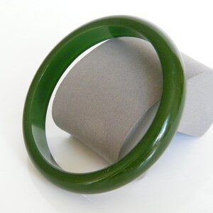 May include: A vibrant, olive green bangle bracelet. The circular bracelet is presented on a grey, cylindrical form against a white backdrop. The bracelet's polished surface reflects light.