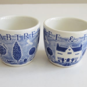 Könnte beinhalten: Zwei weiße Keramik-Eierbecher mit blauen Illustrationen. Ein Becher zeigt Bäume und die Worte "Sweet Home", der andere ein Haus und die Worte "Crabtree & Evelyn". Die Becher haben eine abgerundete Form.