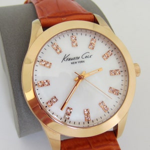 Può includere: Un orologio color oro con quadrante bianco e cinturino in pelle marrone. Il quadrante dell'orologio ha una cornice in oro rosa e presenta il logo Kenneth Cole New York.