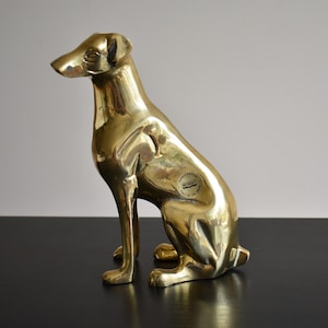 May include: A polished brass figurine of a seated dog with a smooth, reflective surface. The dog is depicted in a sitting position with its head turned slightly to the left. A small circular sticker is visible on the side of the dog's body.