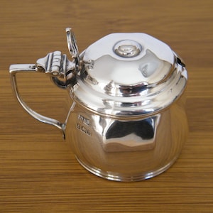 May include: A small, polished silver creamer with a hinged lid and a handle. The creamer has a rounded body and a lid with a small knob. The handle is curved and attached to the side. Likely made of sterling silver, it reflects the light.